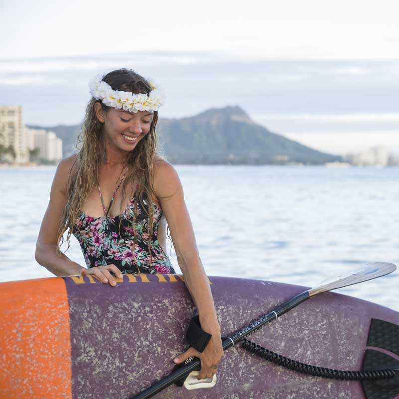 "Pau Hana Surf Supply paddler wearing plumeria lei carrying Carve surf sup and carbon teak paddle with Diamond Head Oahu backdrop"