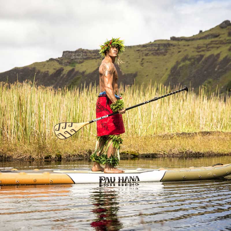 "Pau Hana Surf Supply Endurance Air touring SUP and Endurance fiberglass teak paddle with Hawaiian paddler wearing traditional ti leaf haku lei in scenic mountain waterway"