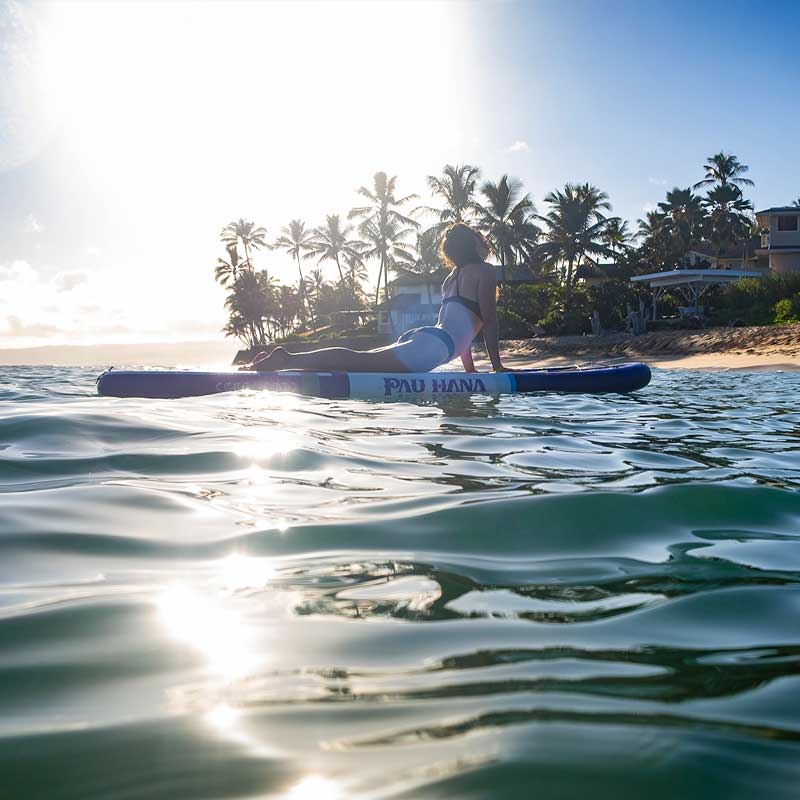 "Pau Hana Surf Supply Moon Mist TPU yoga board action shot - practitioner in cobra pose with tropical Hawaiian sunset backdrop, palms silhouetted"
