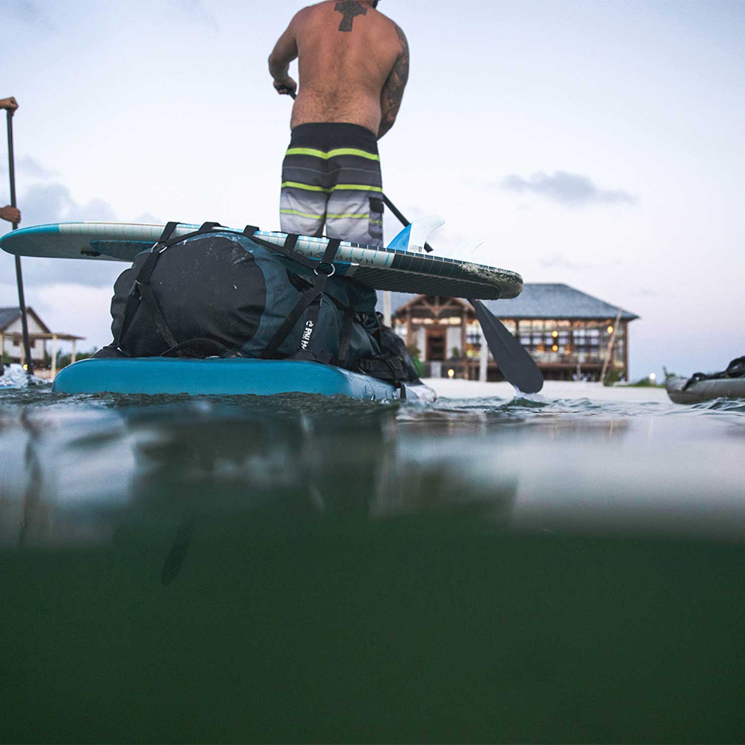 A man paddling a paddleboard loaded up with acessories at dusk towards a hotel on a beach