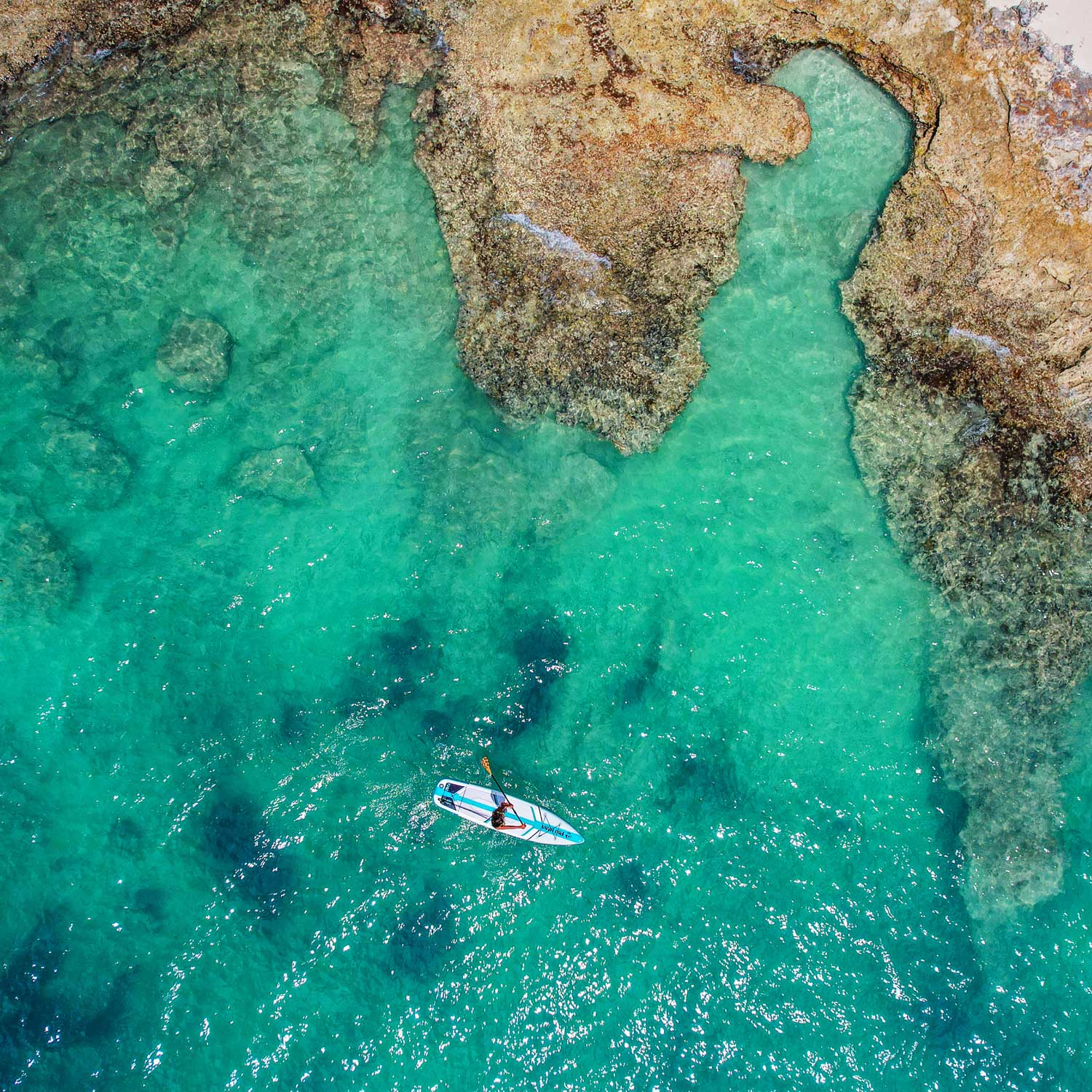 an aerial view of a paddlebaorder paddling past a rocky shoreline on turquoise waters