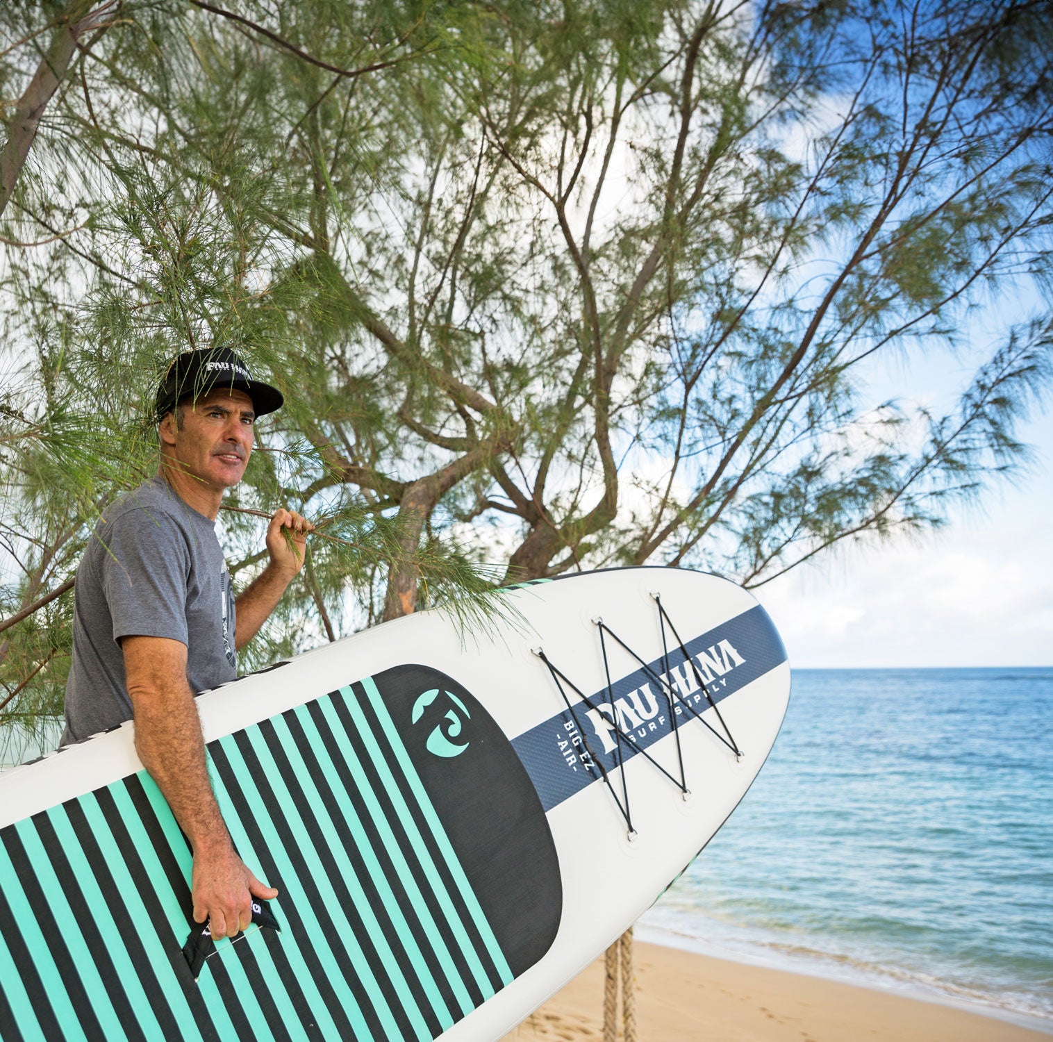 a man holding the big ez air inflatable paddleboard next to the ocean holding onto a tree branch