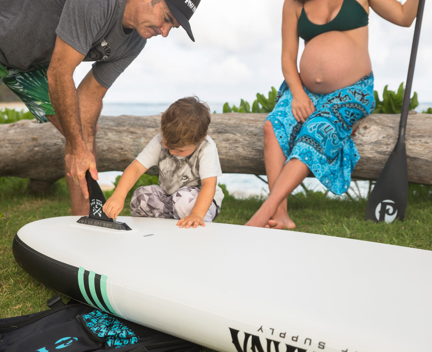 a man helping his son place a fin on a paddleboard with a pregnant lady sitting on a log in the background