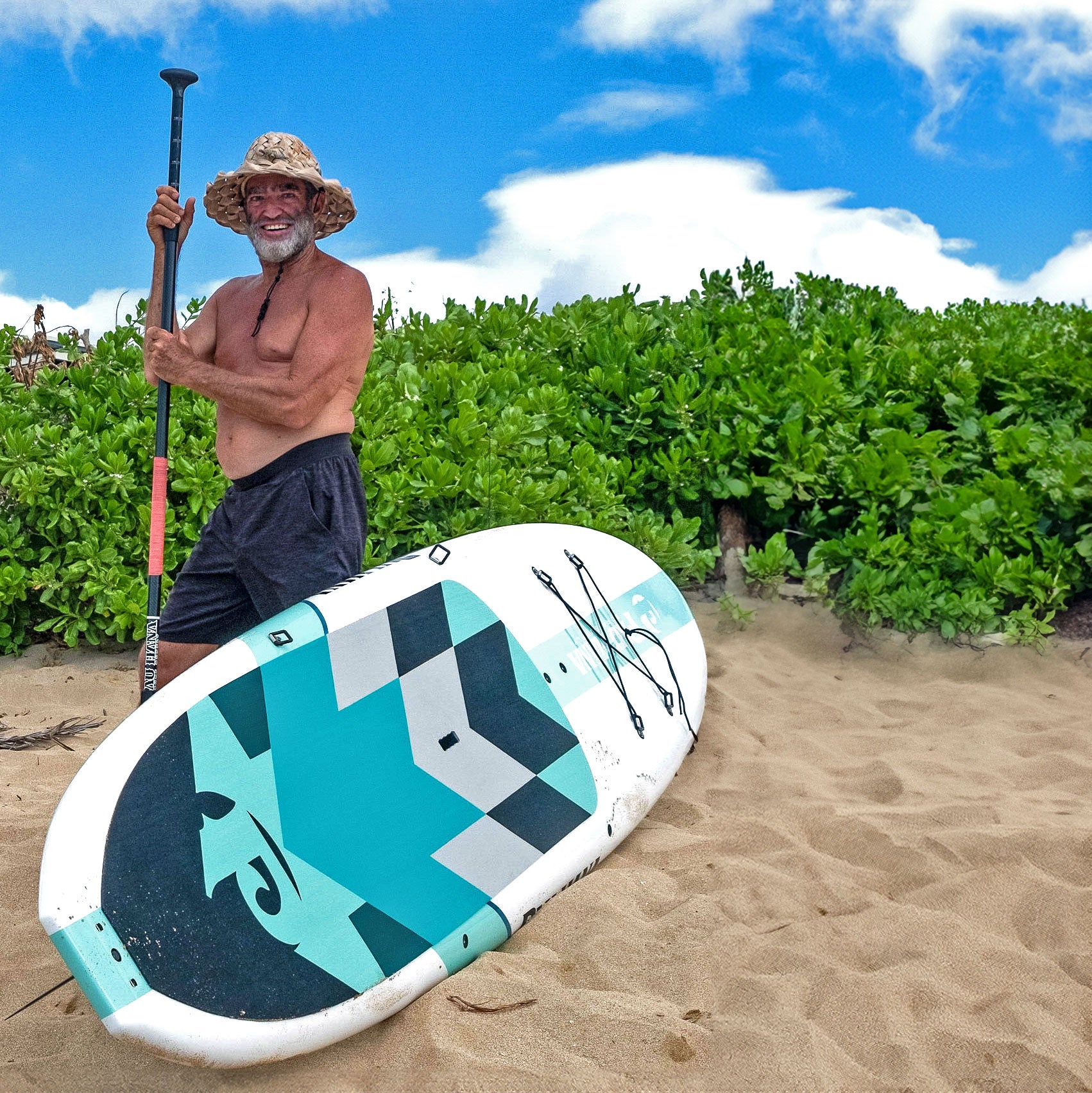 man standing next to a paddleboard on a beach with bushes in the background