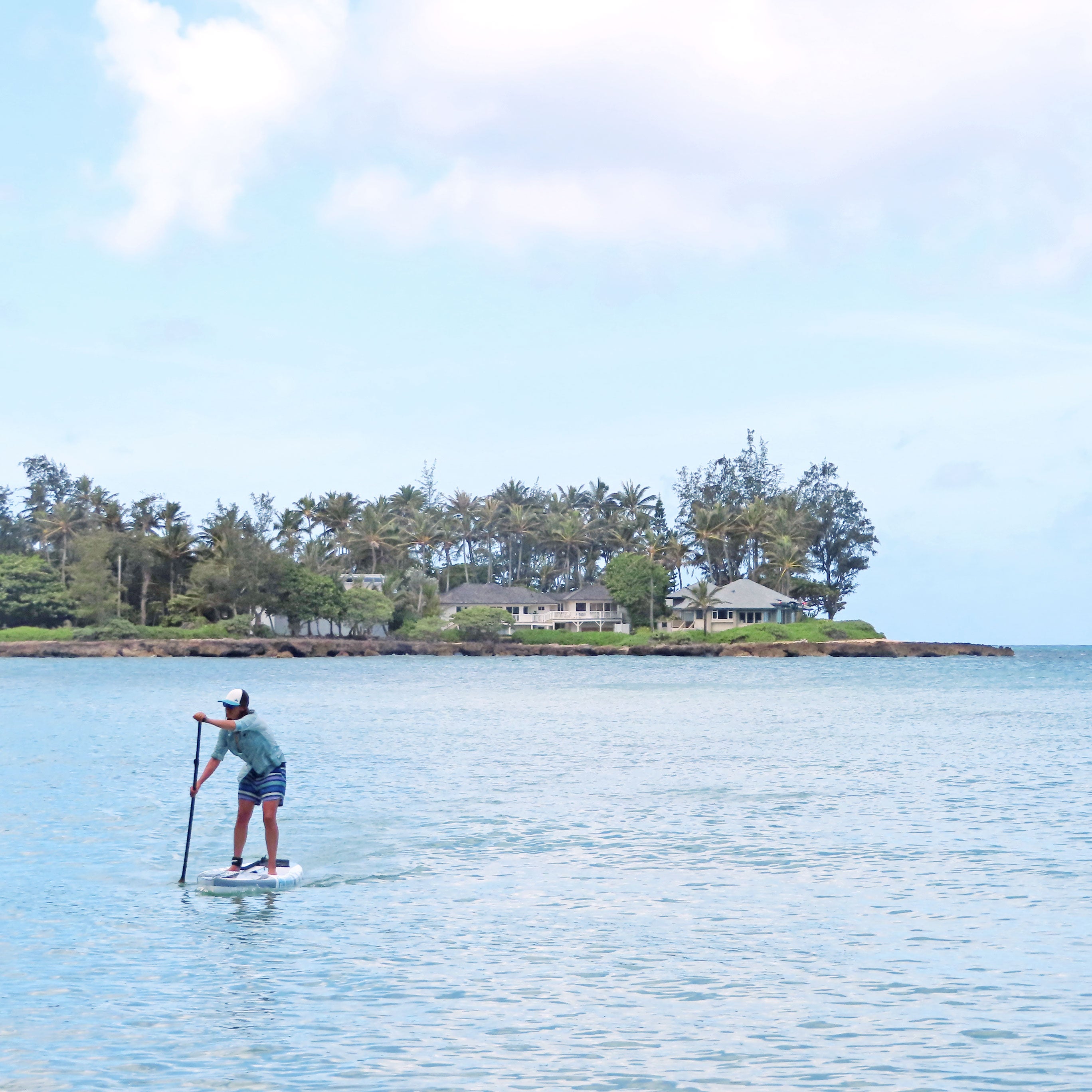 a person paddleboarding on the calm ocean with trees and houses in the background