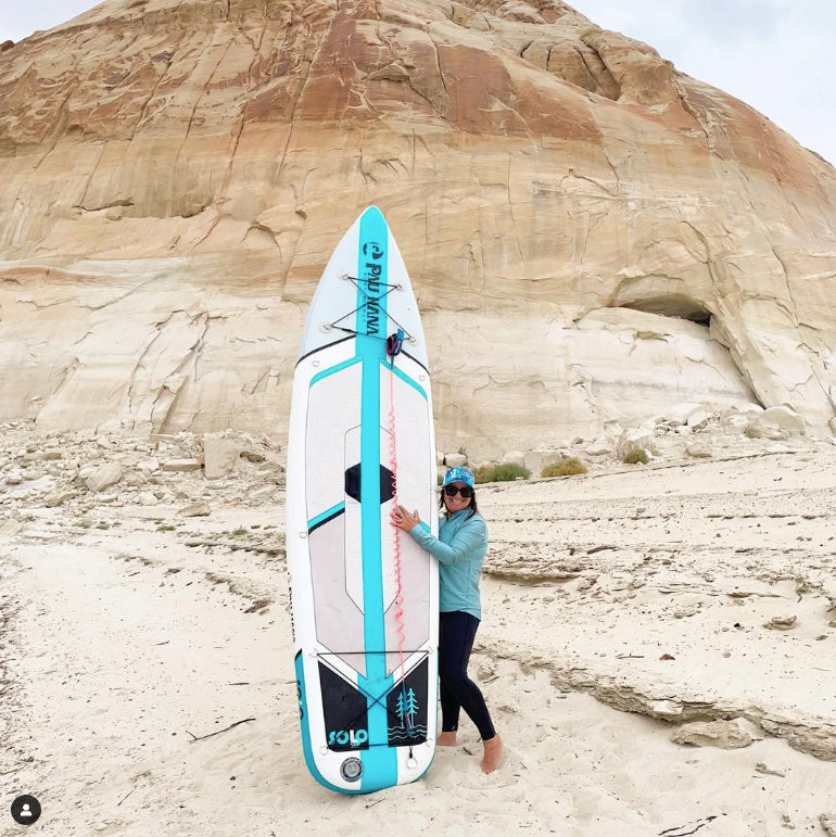 "Pau Hana Surf Supply Solo SUP board displayed in desert landscape, highlighting white and teal colorway with integrated carrying handle"