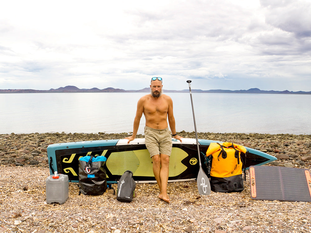 A man stood leaning against a stand up paddleboard surrounded by gear on a pebble beach