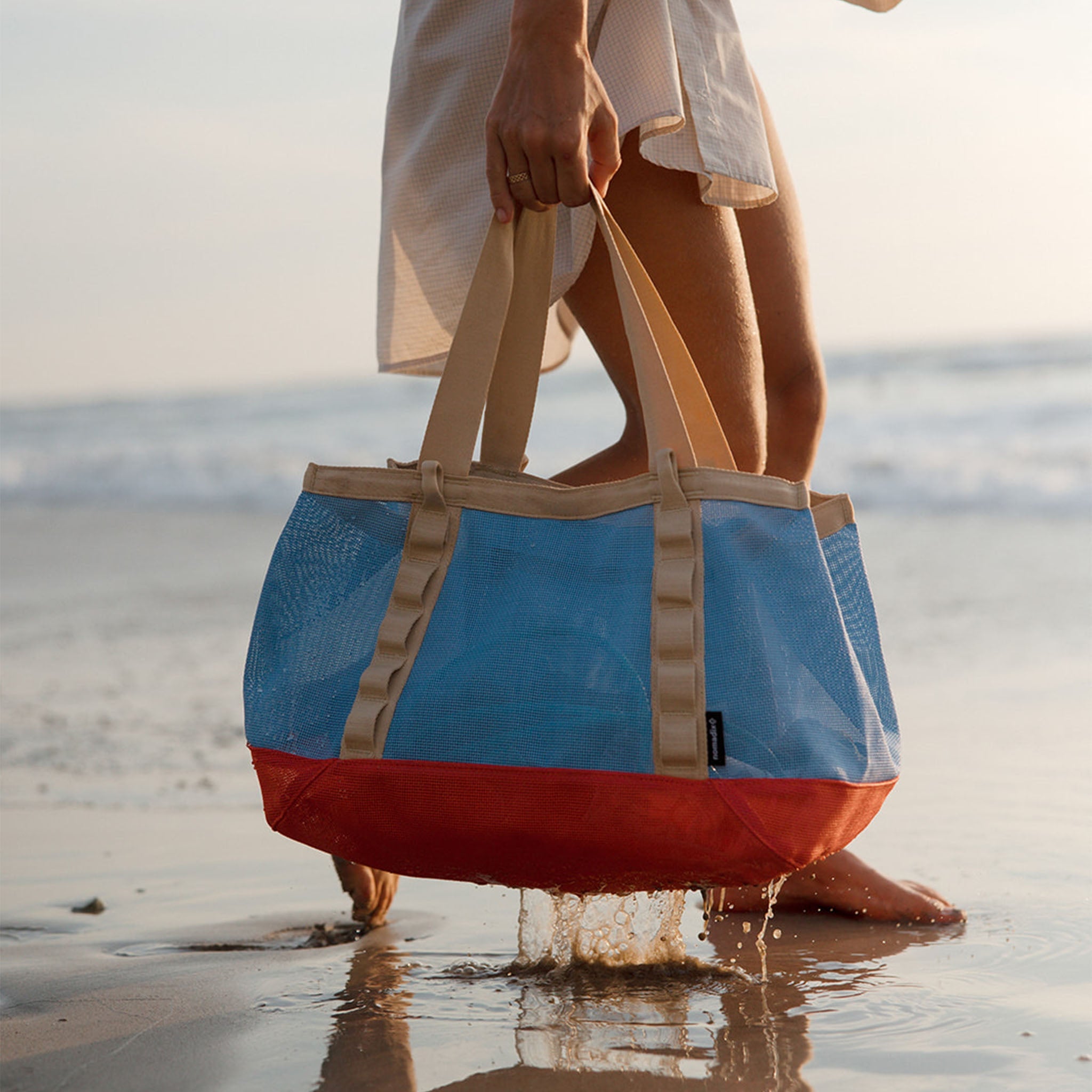 A person on a beach holds a large tote bag with a blue upper section, red lower section, and beige handles and trim.