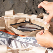 Close-up of a beige mesh tote bag being opened by hands, showing a zippered compartment with a phone inside, set against a dark textured background.
