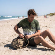 A man sitting on a sandy beach packing a tan canvas backpack, wearing a green and white gradient t-shirt and dark shorts.