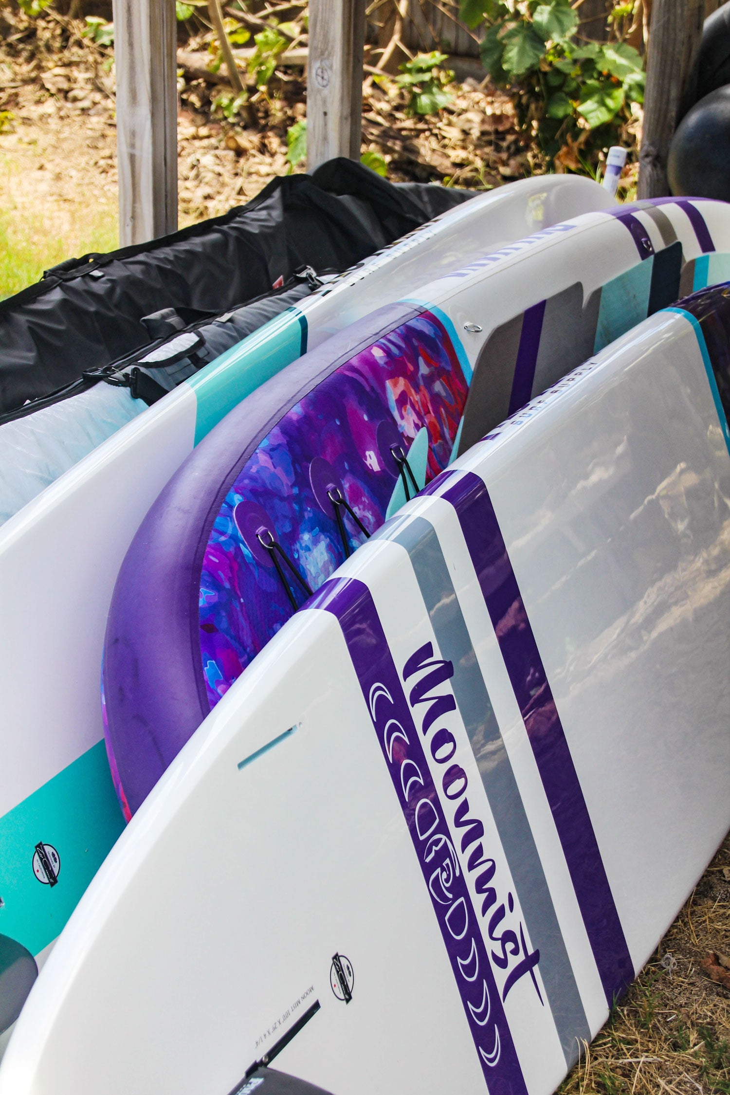 "Pau Hana Surf Supply SUP lineup showing storage with multiple boards - featuring purple cosmic design and turquoise stripes"