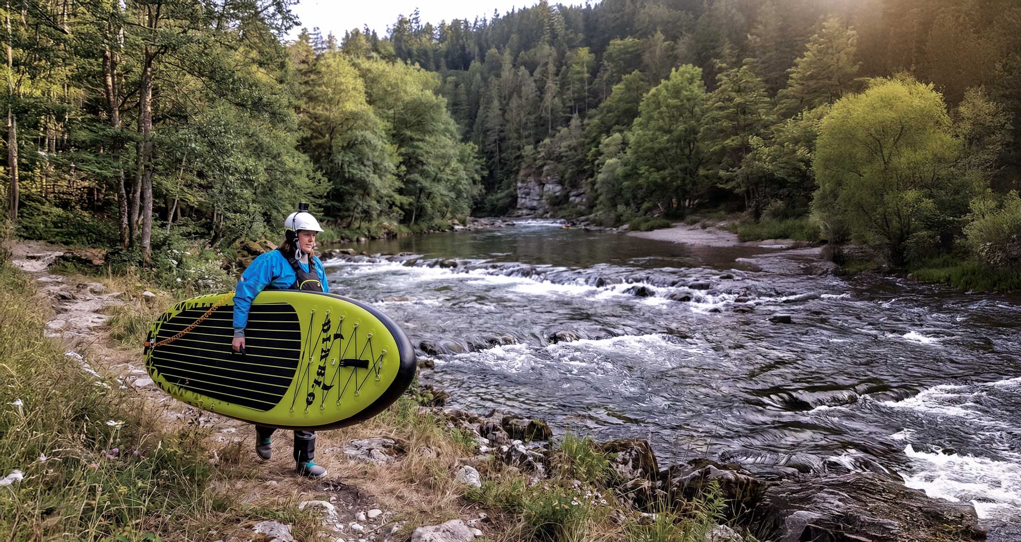 A lady carrying the Pau Hana Diablo Air whitewater SUP by the side of a river with trees and gentle rapids in the background