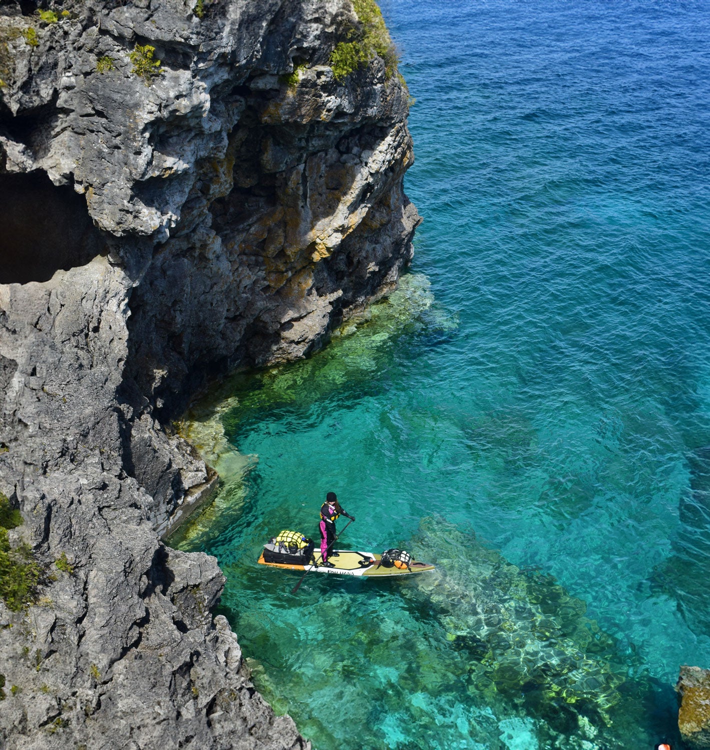 "Pau Hana Surf Supply Endurance touring SUP navigating cold,crystal waters beneath volcanic sea cliffs, showcasing adventure paddling capabilities wearing drysuit and life jacket"