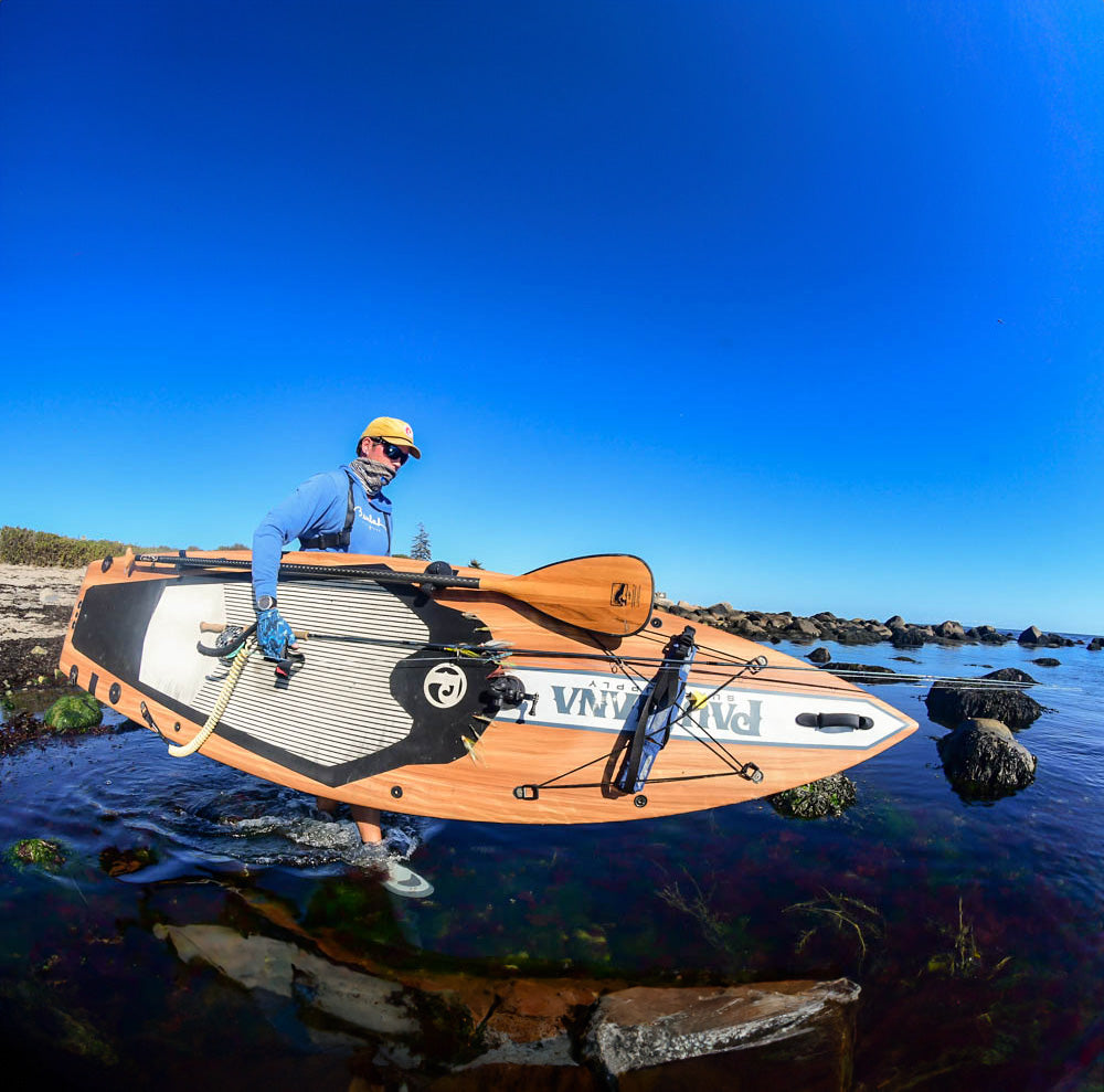 "Pau Hana Surf Supply Endurance VFT paddleboard with fly rod and flies attached to EVA deck pad, showcasing woodgrain deck design against rocky coastal backdrop"