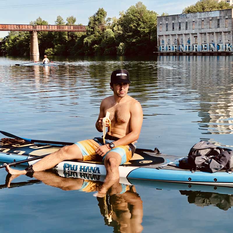 "Pau Hana Surf Supply Endurance XL touring board captured on urban river, paddler resting by industrial waterfront and bridge"