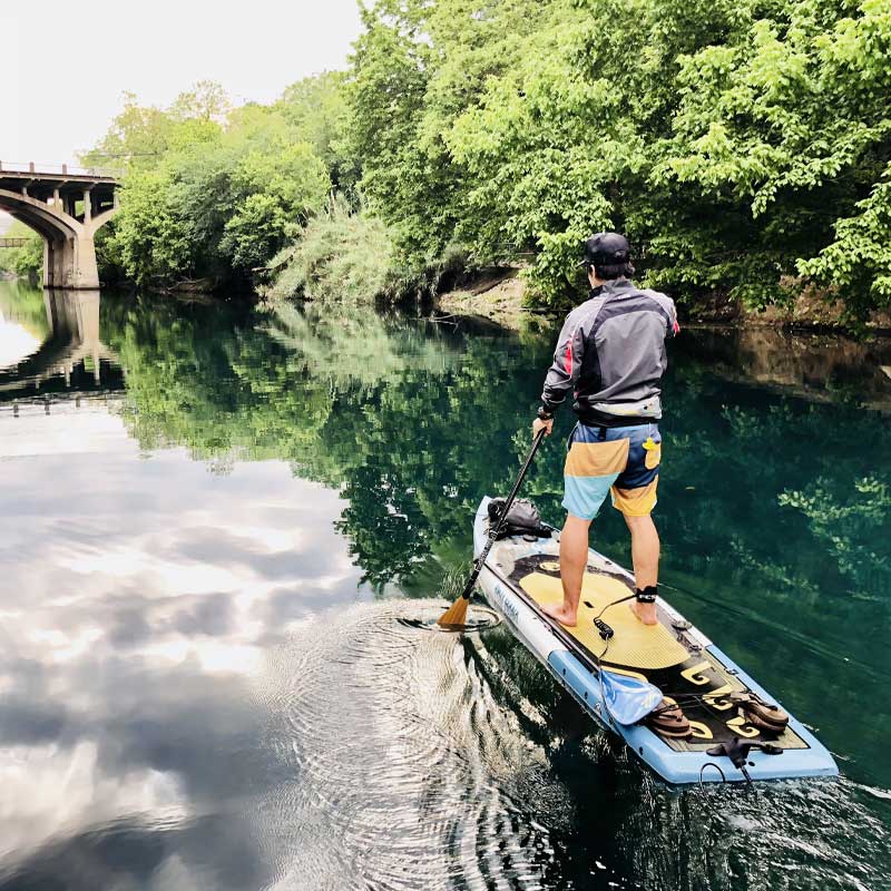 "Pau Hana Surf Supply Endurance XL SUP navigating serene river waters, paddler exploring tree-lined waterway beneath bridge reflections"