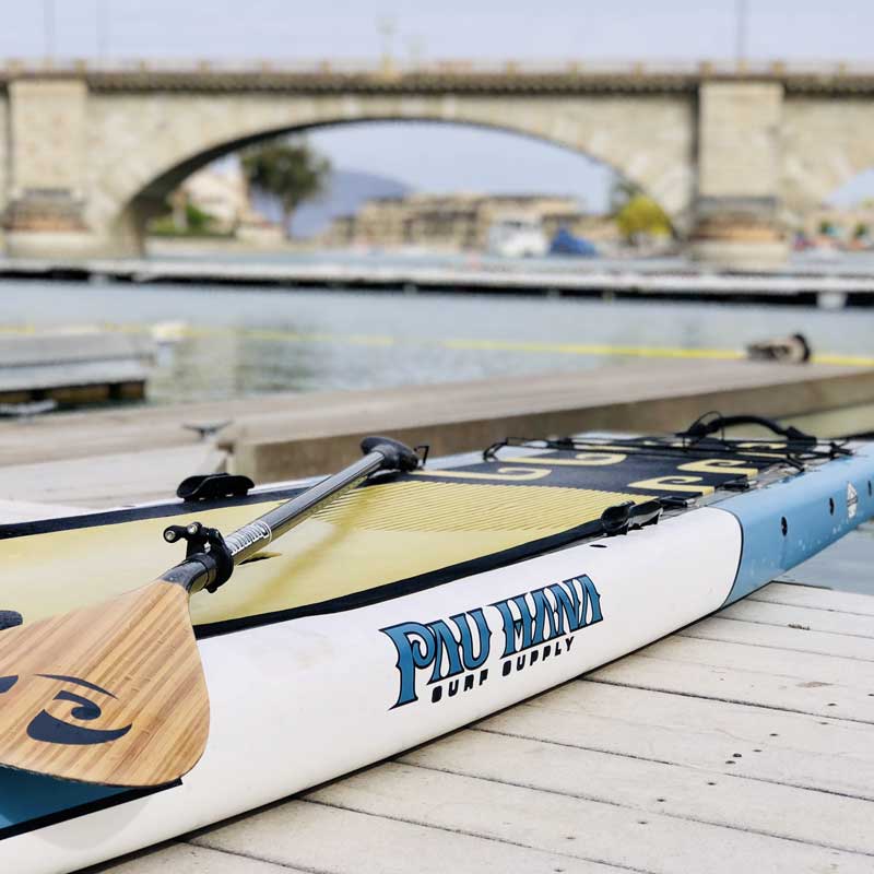 The Pau Hana Surf Supply 12'0" Endurance XL touring paddleboard pulled onto a dock with the carbon teak paddle laying on the EVA deck pad