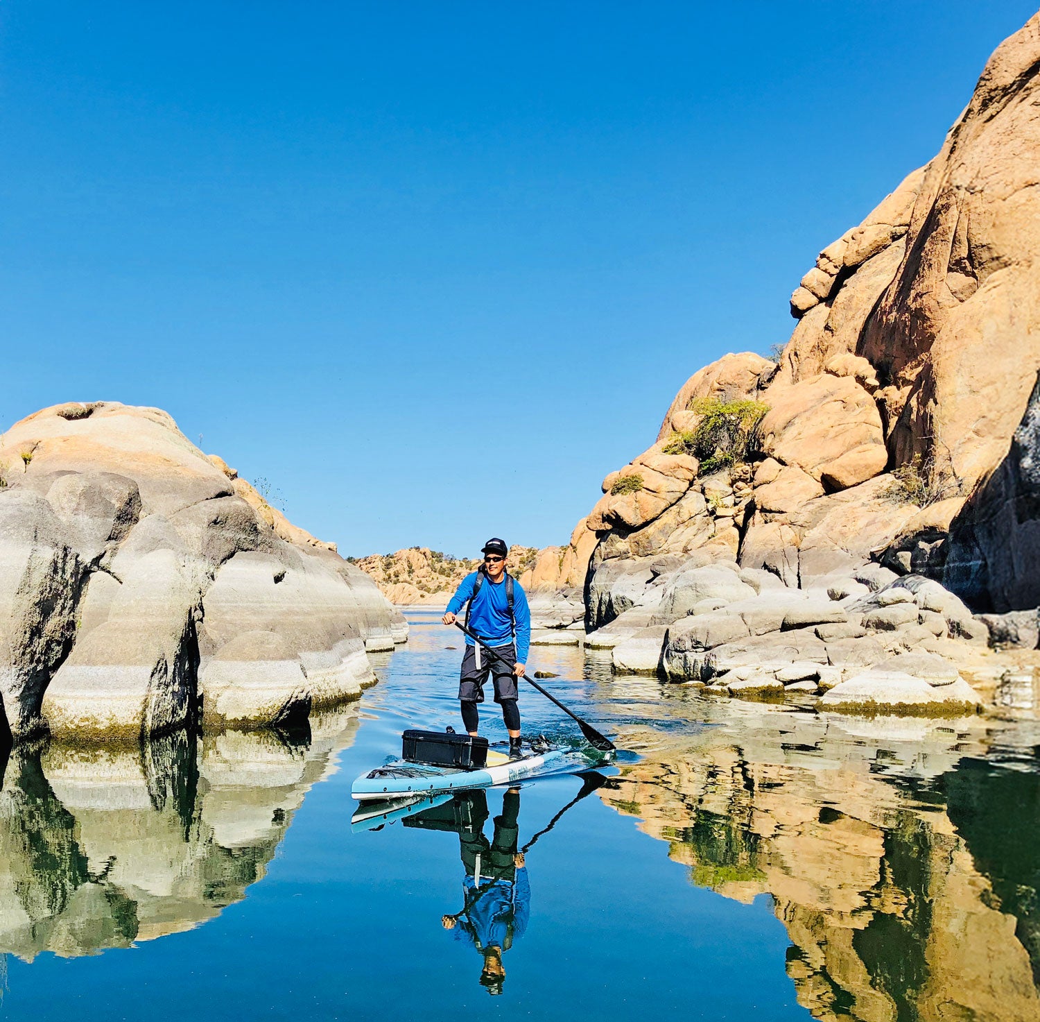 Person paddleboarding on a calm body of water with large rocks and clear blue sky.
