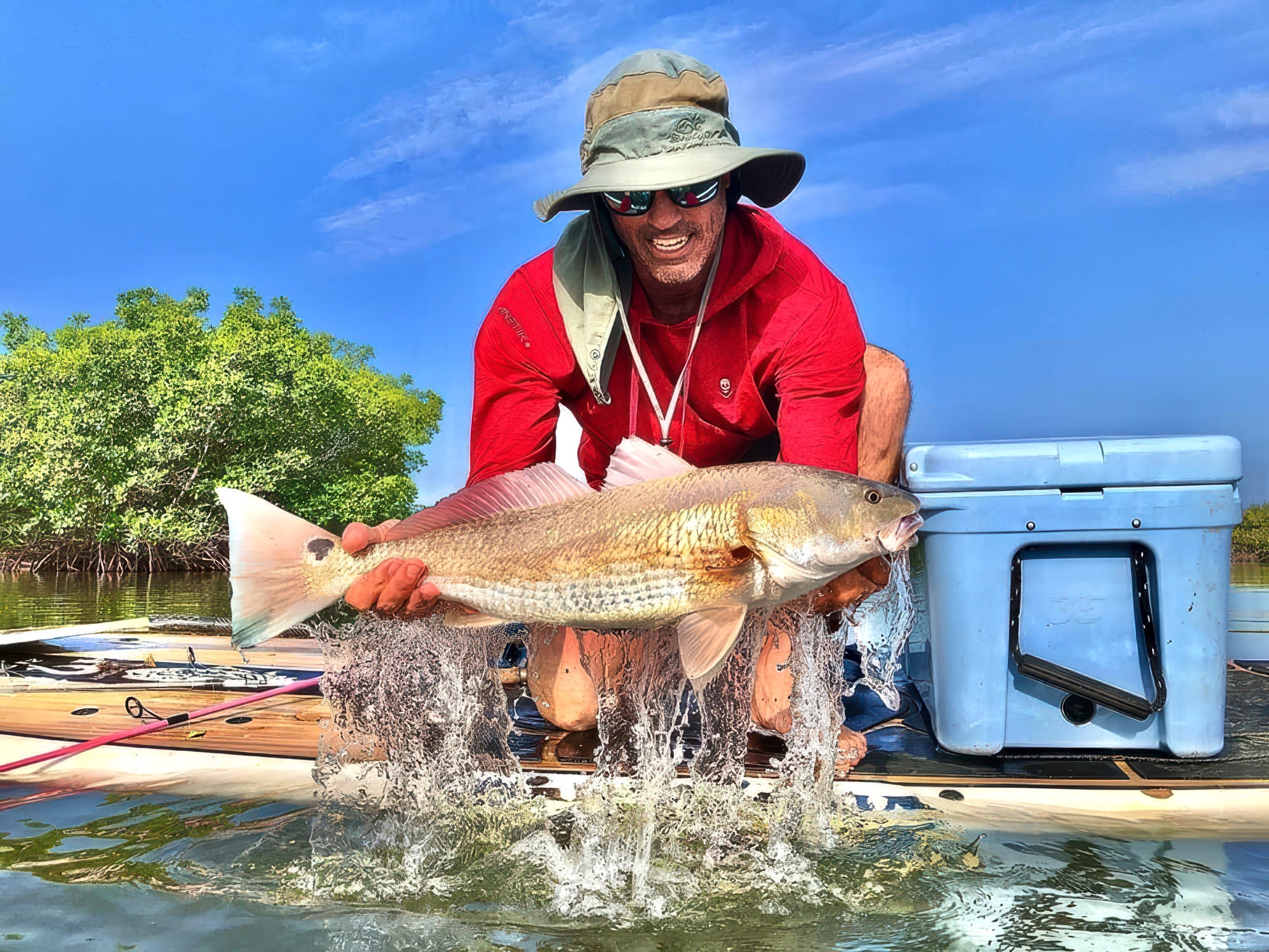 "Successful redfish catch displayed on Pau Hana Surf Supply Angler fishing SUP, water splashing, cooler and tackle visible in Florida mangroves"