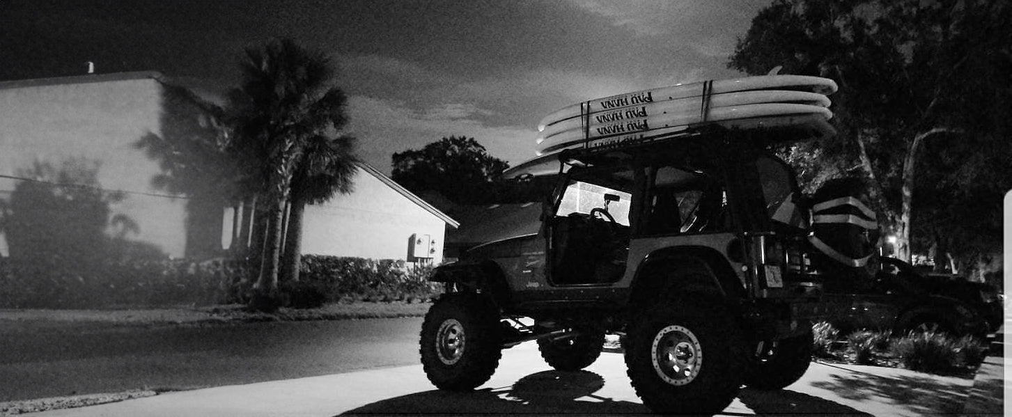 "Modified off-road Jeep with Pau Hana Surf Supply stand up paddleboards mounted on roof rack at dusk, showcasing adventure-ready SUP transport"