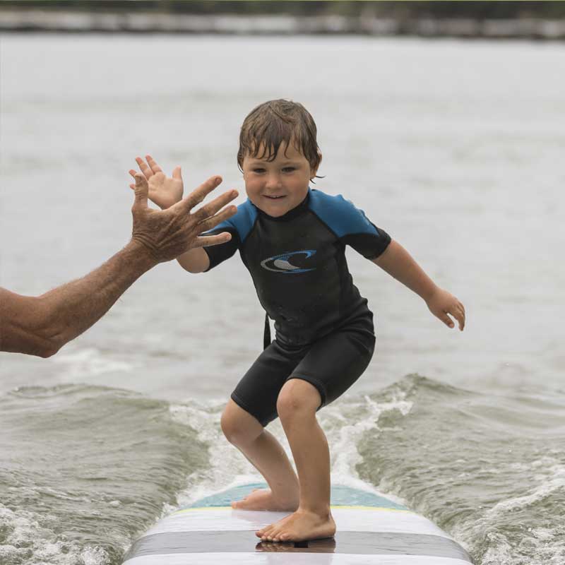 "Young surfer celebrating high-five while riding Pau Hana Surf Supply Grom X kids SUP, showing confident stance in small waves"