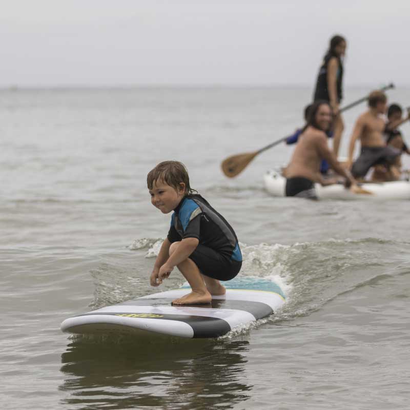 "Young surfer catching wave on Pau Hana Surf Supply Grom X kids SUP, with family paddling Oahu Nui in background"