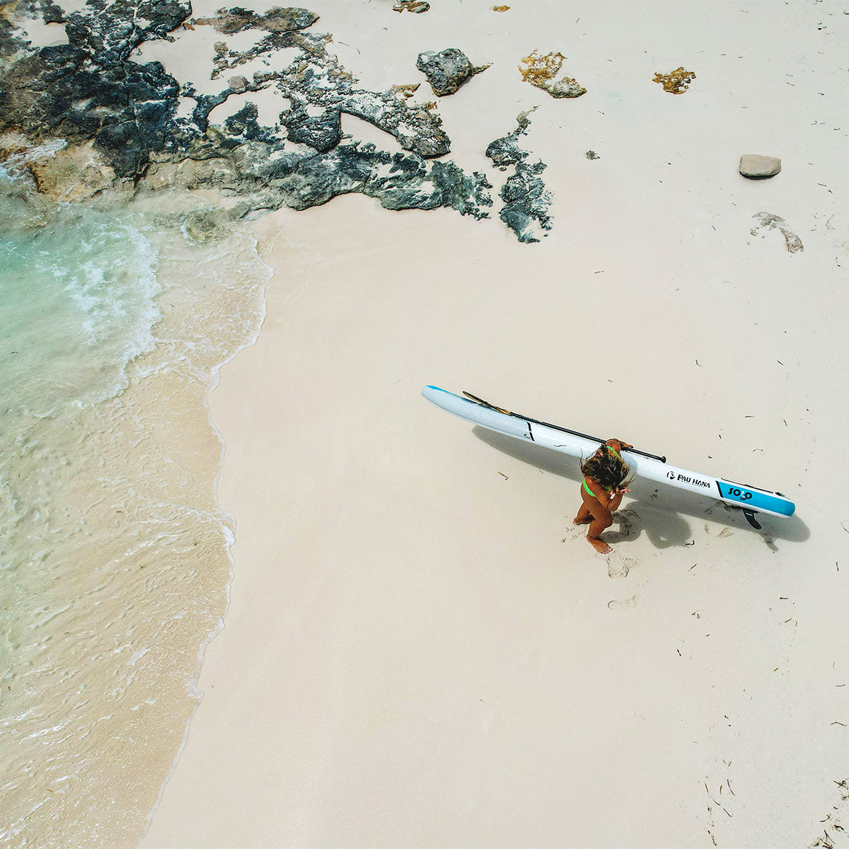 "Aerial view of paddler carrying Pau Hana Surf Supply 10'10" Solo SUP backcountry lightweight paddleboard on pristine white sand beach with turquoise water and volcanic rocks"