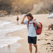 A man walking on a sandy beach wearing a light blue t-shirt, dark shorts, and carrying a red backpack.