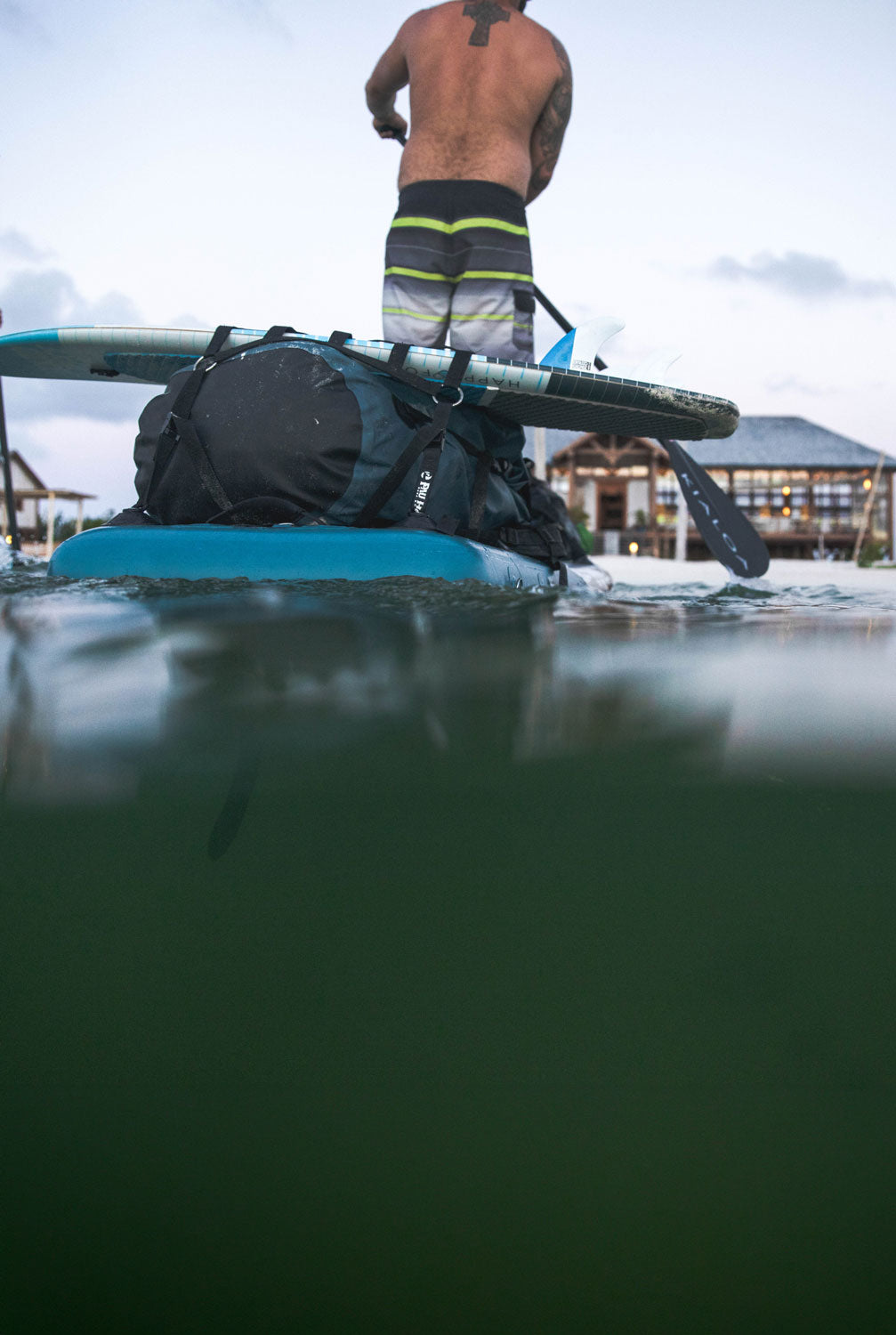 "Split-shot waterline view of Pau Hana Surf Supply Endurance Air touring SUP with mounted kitesurf and gear bag in tropical waters"