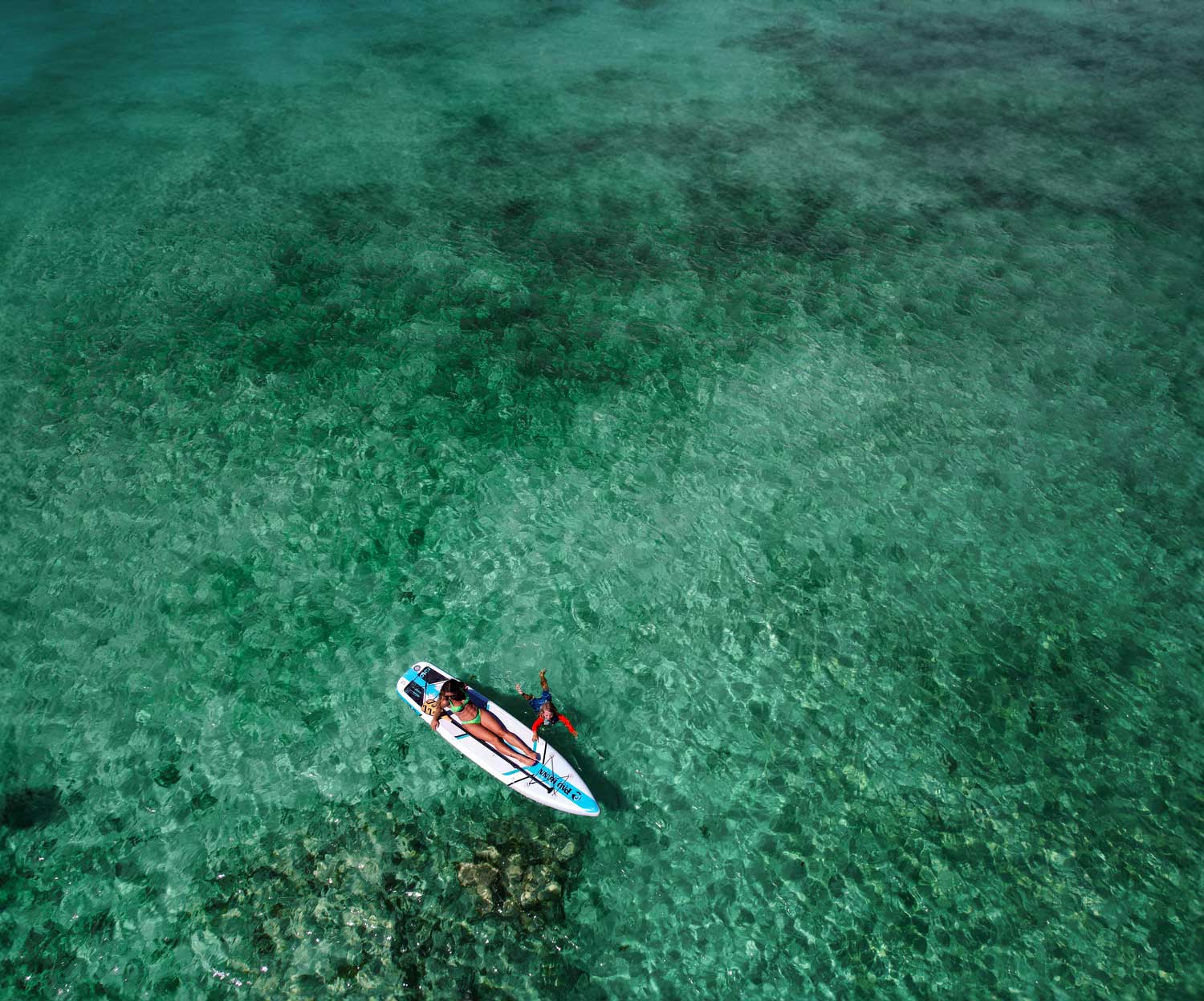 "Pau Hana Surf Supply Solo SUP floating over coral reef - aerial view showing crystal-clear Caribbean waters and inflatable backpacking touring board design"