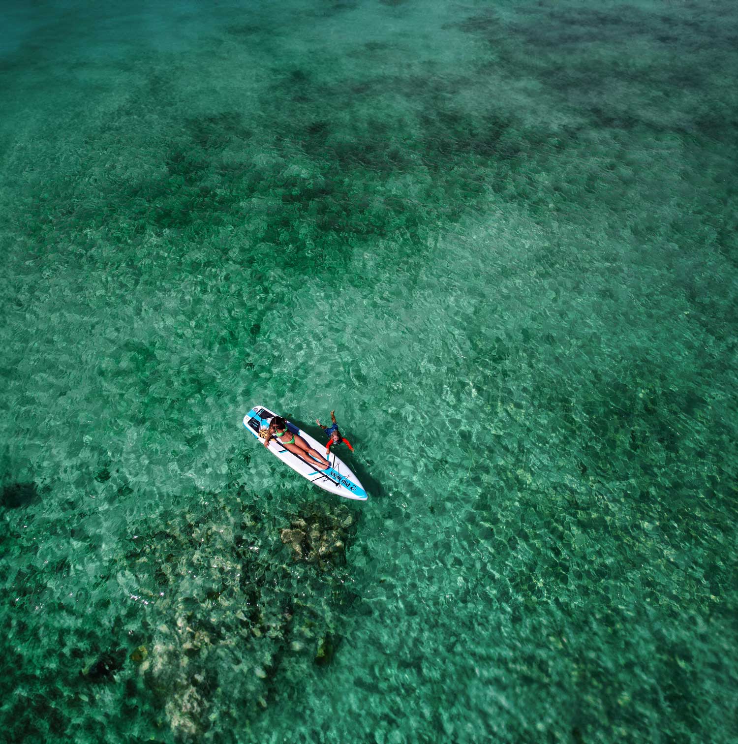 "Pau Hana Surf Supply Solo SUP floating over coral reef - aerial view showing crystal-clear Caribbean waters and inflatable backpacking touring board design"