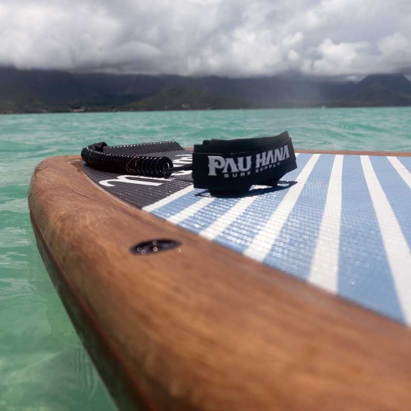 Pau Hana Surf Supply Malibu Tour VFT stand up paddleboard with wood rail detail and soft grip deck pad floating on turquoise Hawaiian waters against mountain backdrop