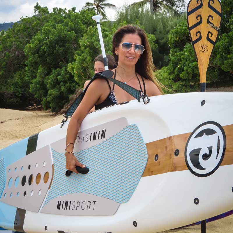 "Woman carrying a Pau Hana Surf Supply 10'10" Mini Sport teal and white with the Kamba Strap holding an Endurance fiberglass teak paddle