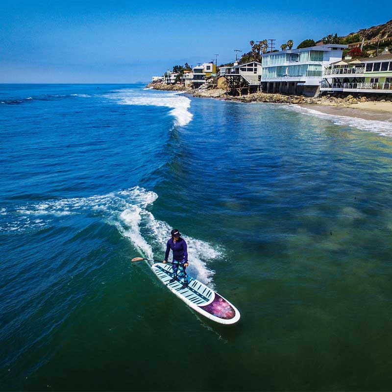 "Aerial view of surfer riding Pau Hana Surf Supply Moon Mist Sup SUP surf and yoga board on ocean wave along Malibu beachfront with modern coastal homes"