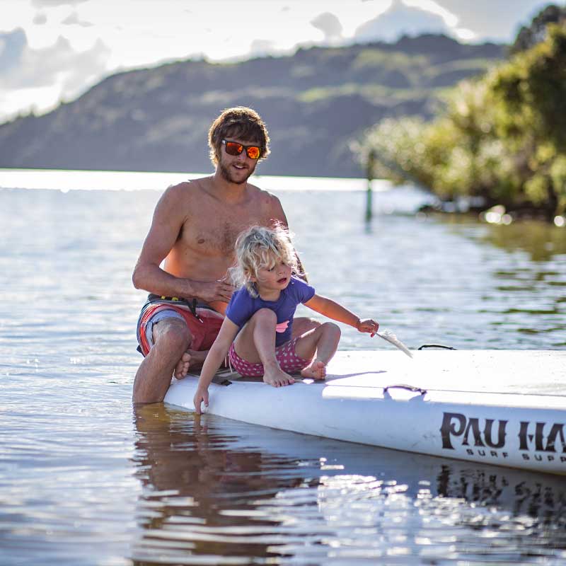 A father and child on the Pau Hana Surf Supply Oahu Nui giant inflatable family paddleboard on a lake with trees in the background