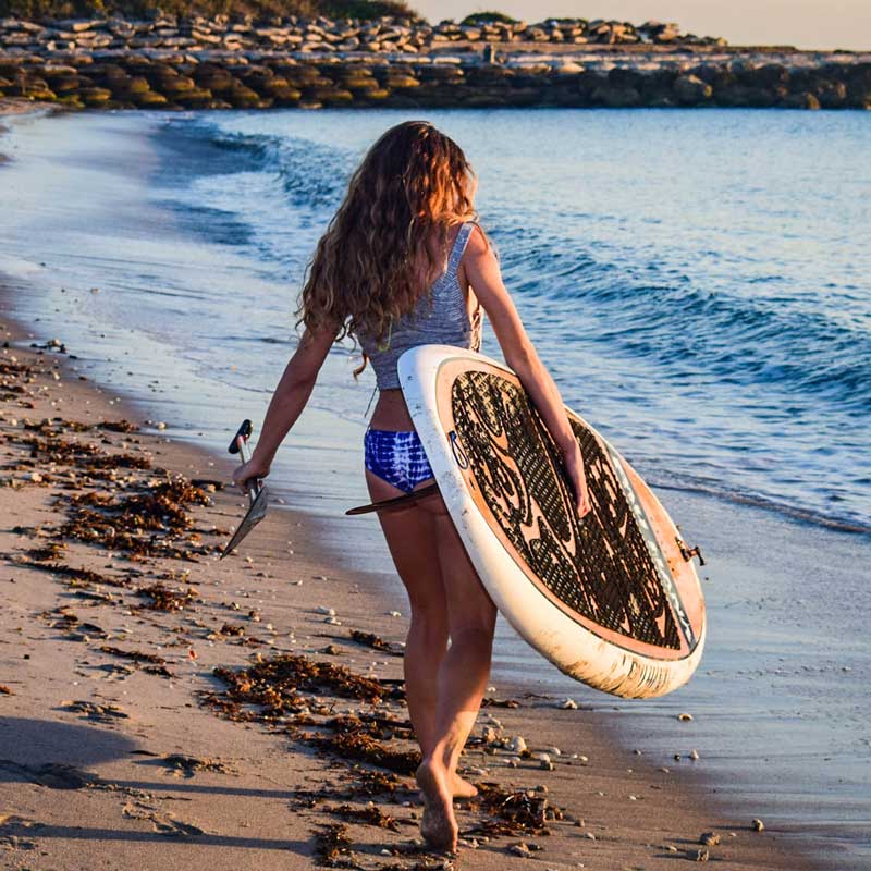 &quot;Woman carrying a Pau Hana Surf Supply 10&quot;0&quot; Oahu SUP with the paddle walking towards the ocean&quot;