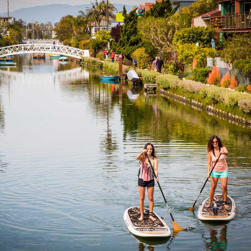 "Two paddlers cruising Venice Canal on the Pau Hana Surf Supply 10'0" Oahu paddle board in white with carbon teak paddles, white bridge backdrop"