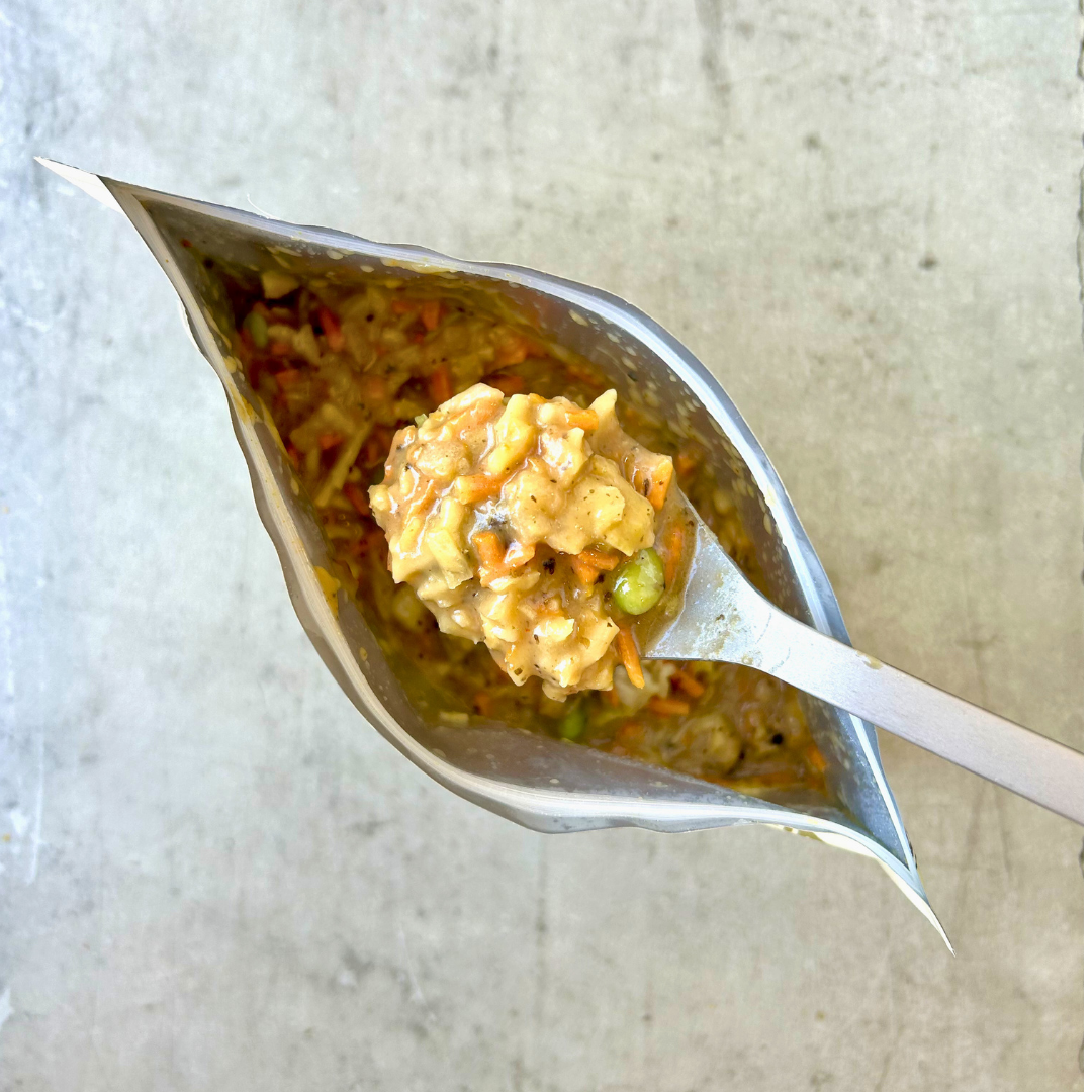 A close-up shot of creamy, chunky orange-colored rice or oatmeal with vegetables, held on a metal spoon inside a silver foil pouch against a light gray textured background.
