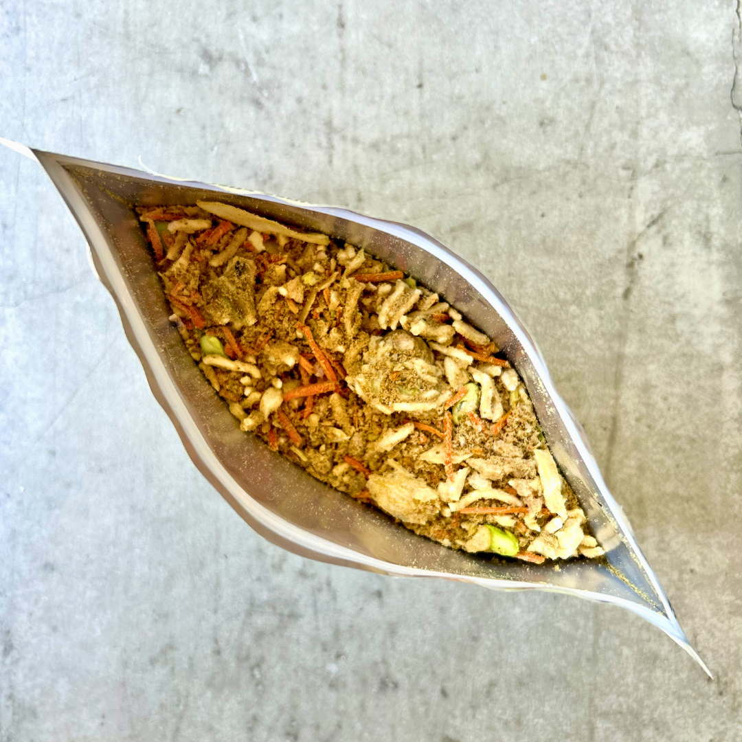 An open silver foil pouch filled with a loose herbal or pet chow mix, photographed from above on a light gray textured surface.