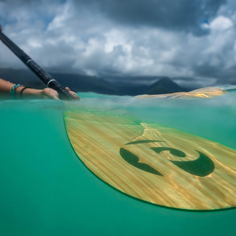 "Pau Hana 3-piece carbon teak paddle split-shot underwater view in Hawaiian turquoise waters, showcasing signature tribal logo"