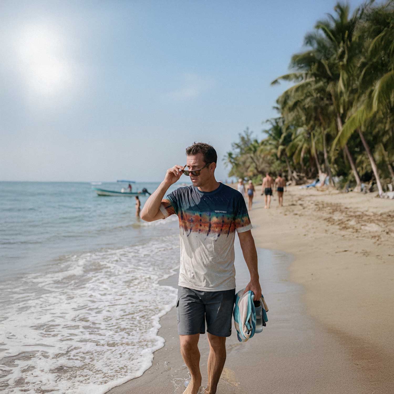 A man walking on a sandy beach wearing a white and blue tie-dye t-shirt, dark gray shorts, and sunglasses, holding a striped towel. Palm trees and the ocean are visible in the background.