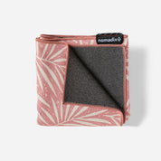 A folded Nomadix travel blanket with a dusty pink and cream palm leaf pattern on one side and a dark gray fleece lining on the other, shown on a light gray background.