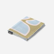 A folded textile item, likely a blanket or towel, featuring an abstract pattern in light blue, pale pink, and a muted olive-beige color, with a small black tag on the edge.