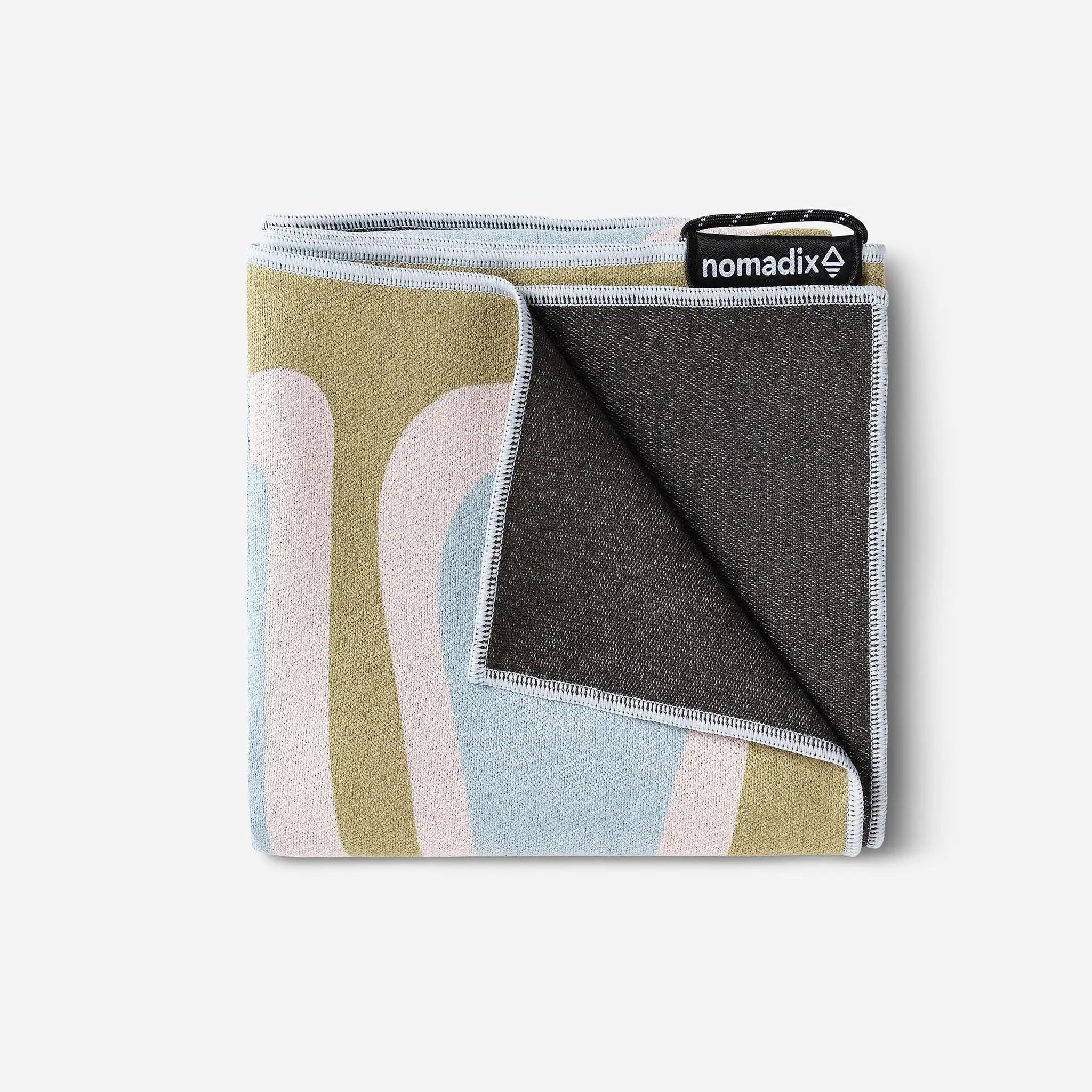 A folded Nomadix blanket with a multicolor abstract pattern in olive green, light blue, and pale pink, showing a dark gray fleece backing and a black brand tag.