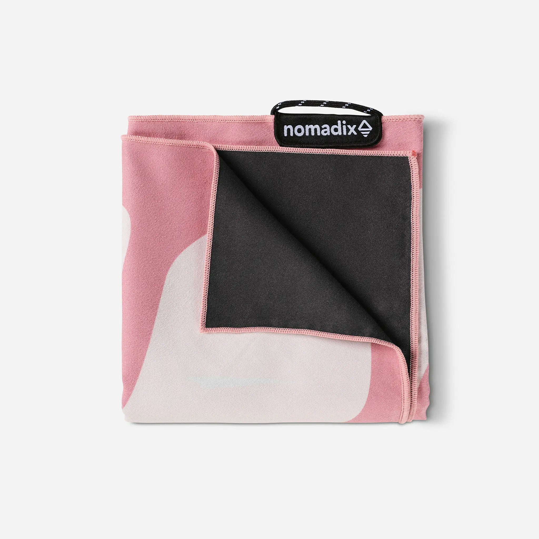 A folded Nomadix microfiber blanket in a pink, white, and black abstract pattern, shown on a light gray background with its black carrying strap visible at the top.