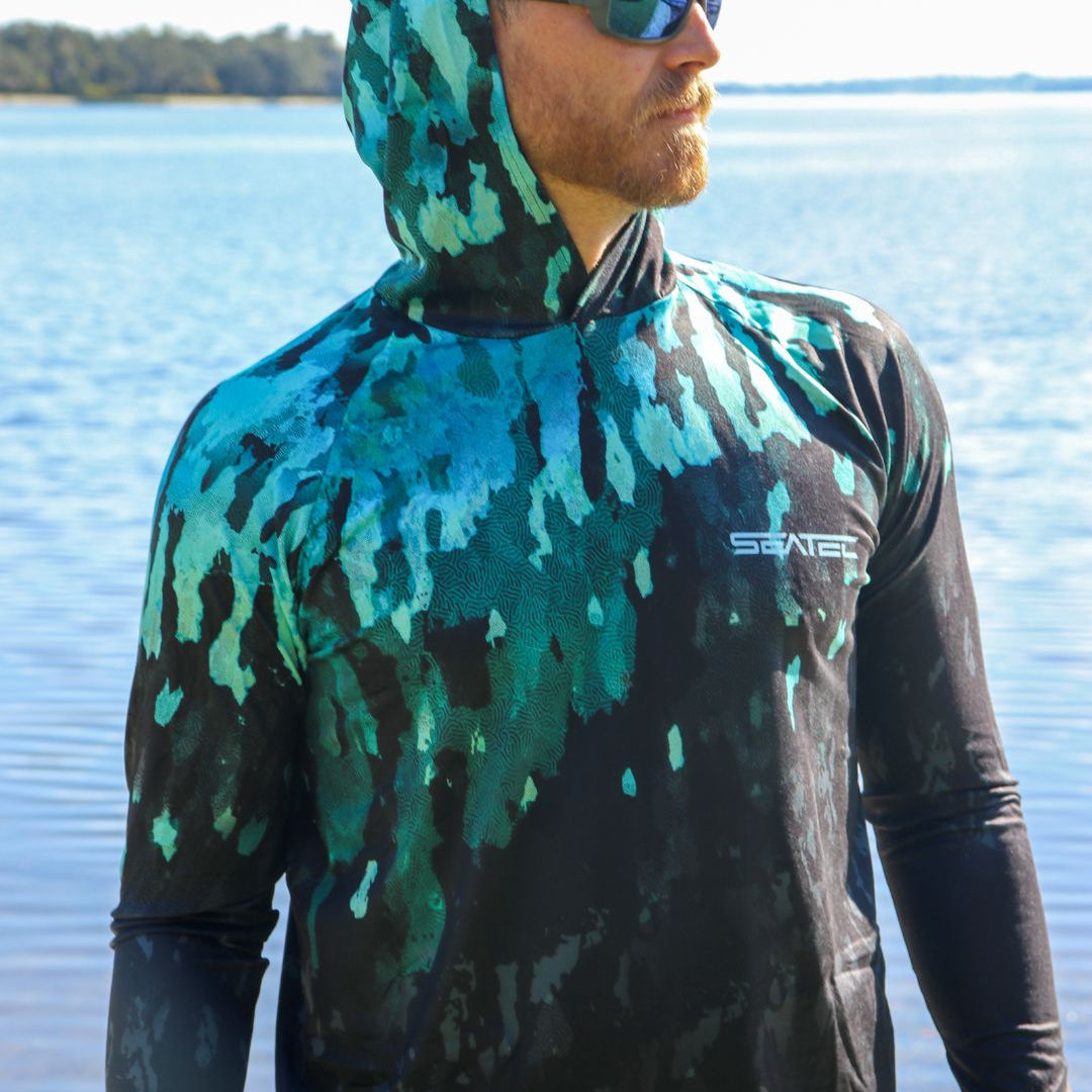 Man wearing a camouflage hoodie with a brand logo, standing by a body of water.