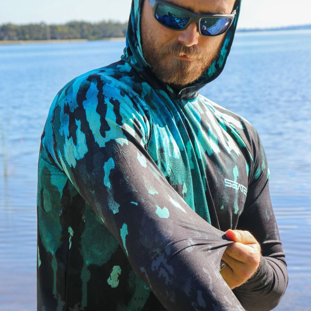 Man wearing a camouflage hoodie and sunglasses by a body of water