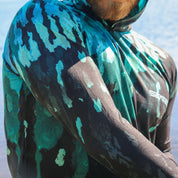 Person wearing a camouflage hooded shirt with a brand logo, standing by a body of water.