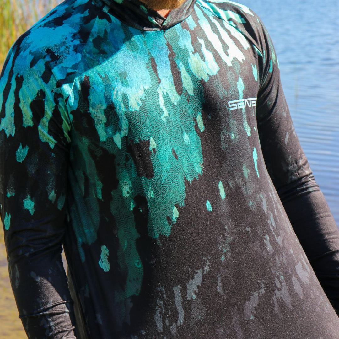 Person wearing a camouflage-patterned shirt with a brand logo, standing by a body of water.
