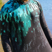 Person wearing a camouflage-patterned shirt with a brand logo, standing by a body of water.